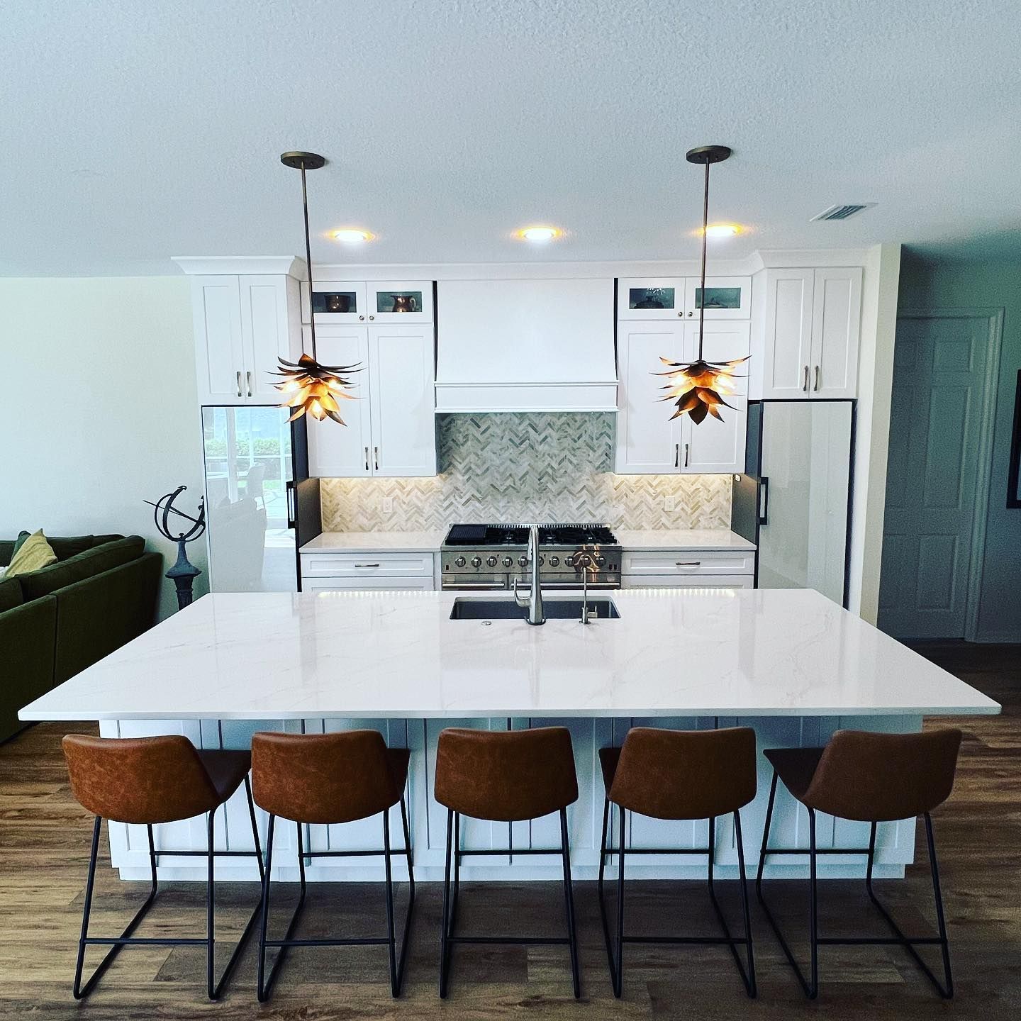 Modern white kitchen with island, brown stools, pendant lights, and stainless steel appliances.