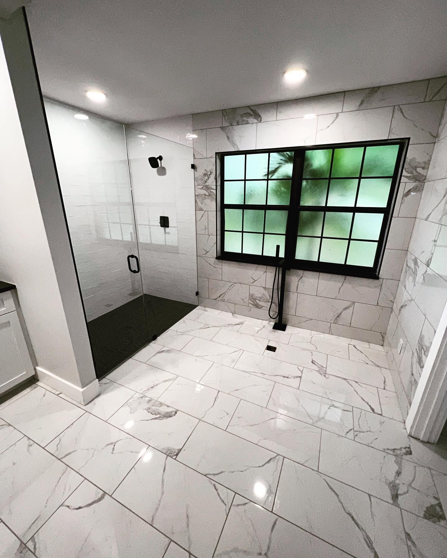 Modern bathroom with white marble tile, black shower fixtures, and a window with a green outdoor view.