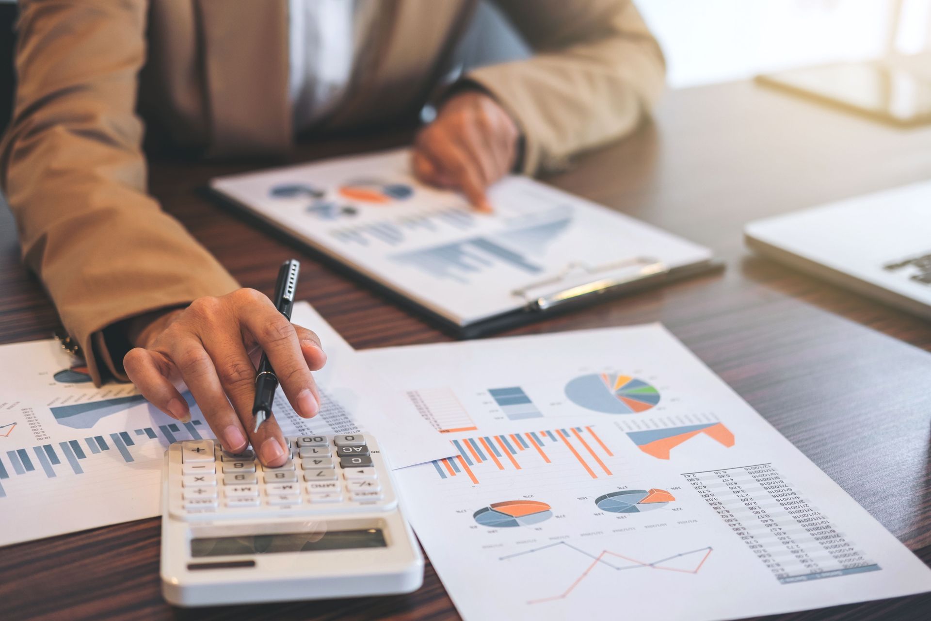 Person in blazer using calculator, reviewing financial reports with charts and graphs on a wooden table.