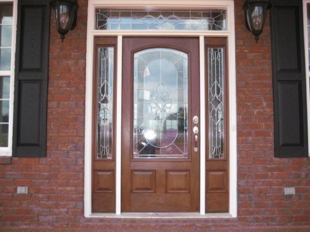 Brick house front door with decorative glass panels and sidelights, framed by black shutters.