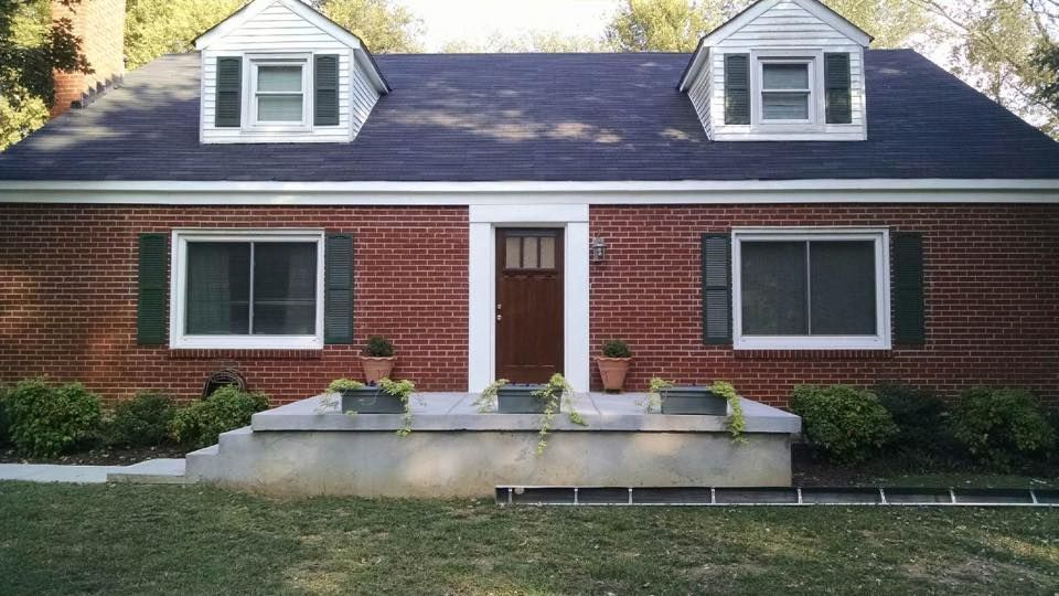 Brick house with white dormers, brown door, and front stone planter beds under a dark roof