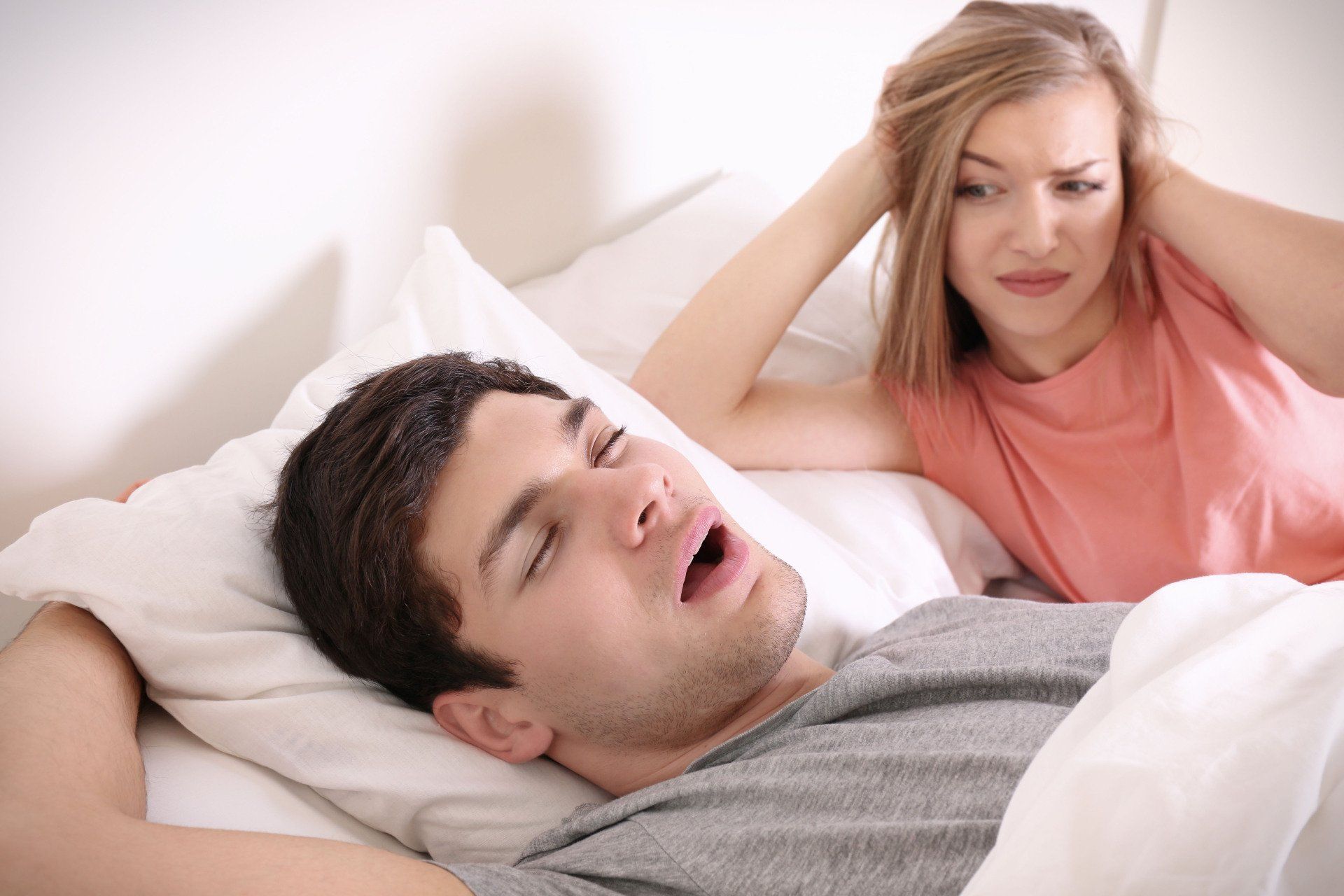 A woman is covering her ears while a man is snoring in bed.