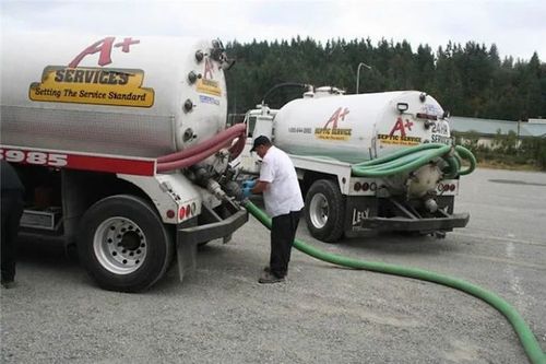 Workers by two white service trucks, one filling a green hose in a gravel lot.