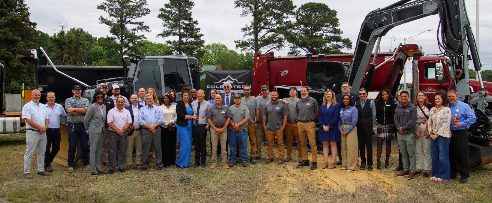 Group posing outdoors beside excavators and construction trucks at a work site