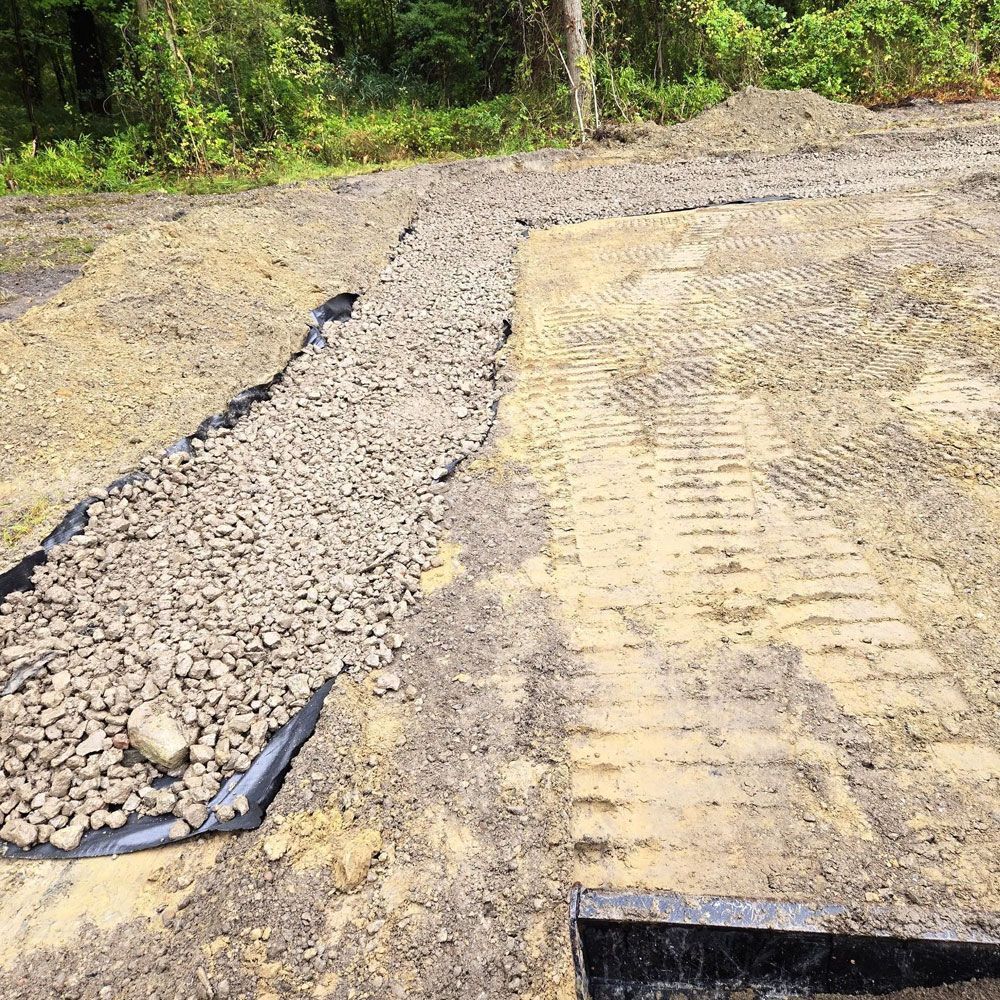 A gravel-filled drainage trench lined with black fabric runs through a dirt construction site with tire tracks.