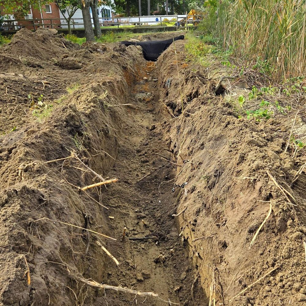A narrow dirt trench dug into the ground at an outdoor construction site, showing layered soil and roots.