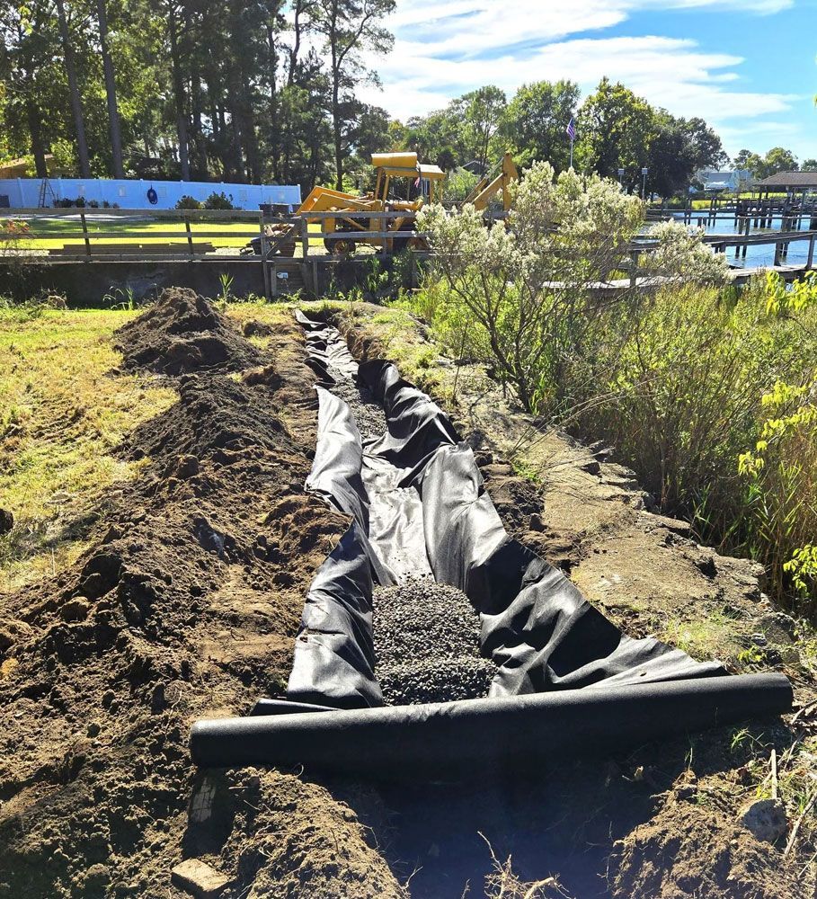 A trench lined with black landscape fabric and partially filled with gravel for a drainage project near a waterfront.