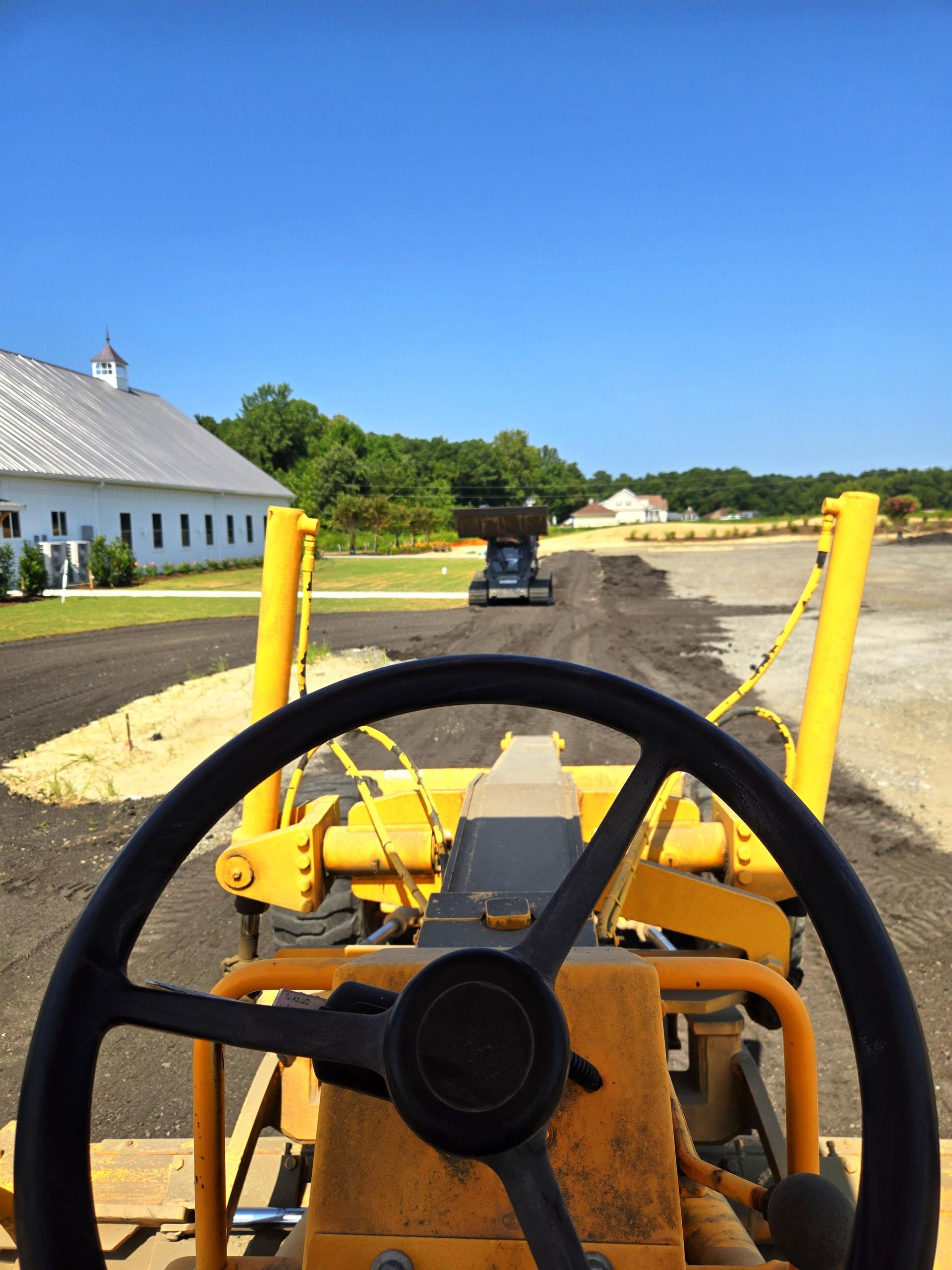 View from the operator's seat of yellow construction equipment leveling a dirt path toward a white building.