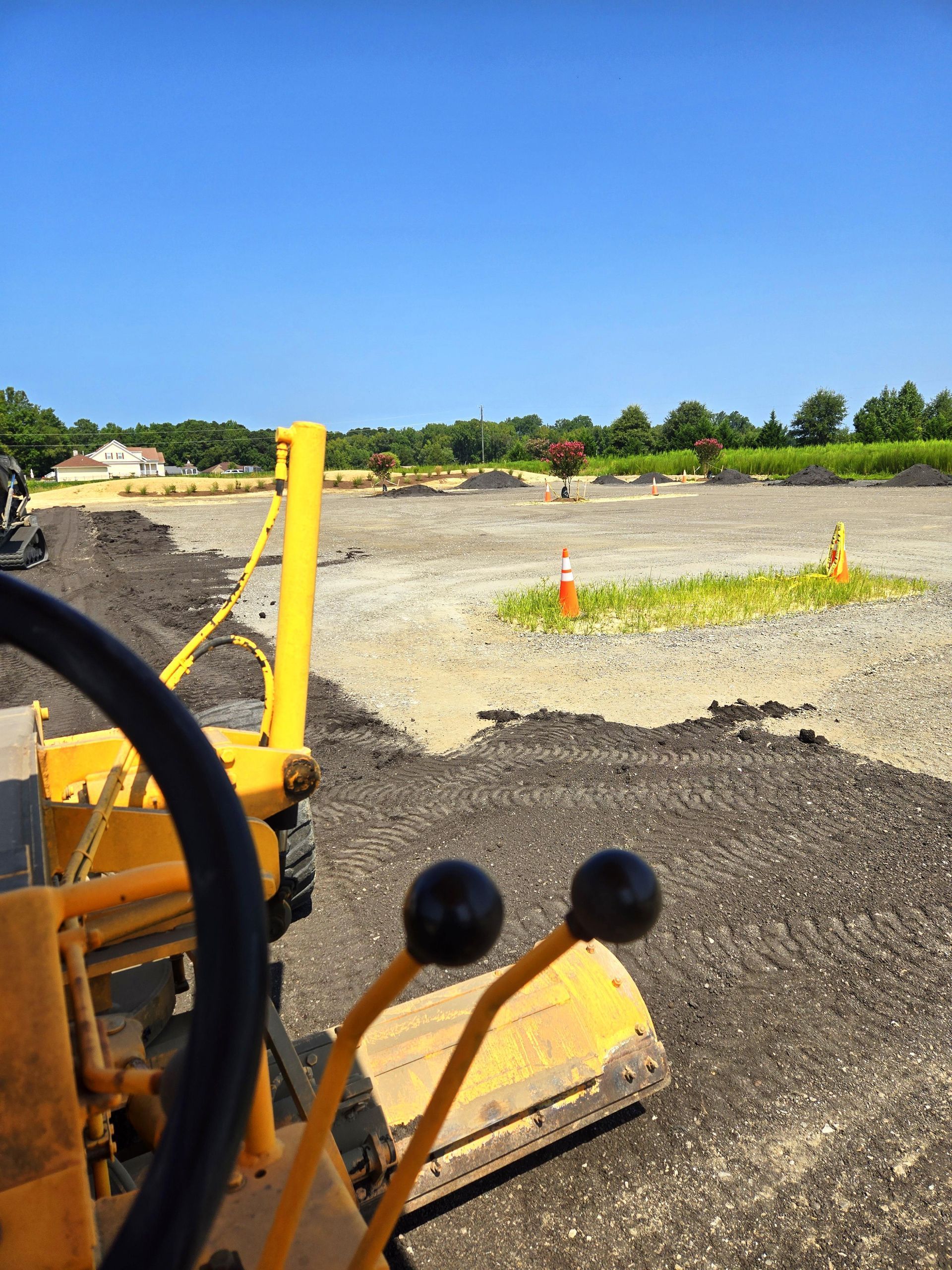 A view from the cab of a yellow construction vehicle overlooking a cleared dirt lot under a clear blue sky.