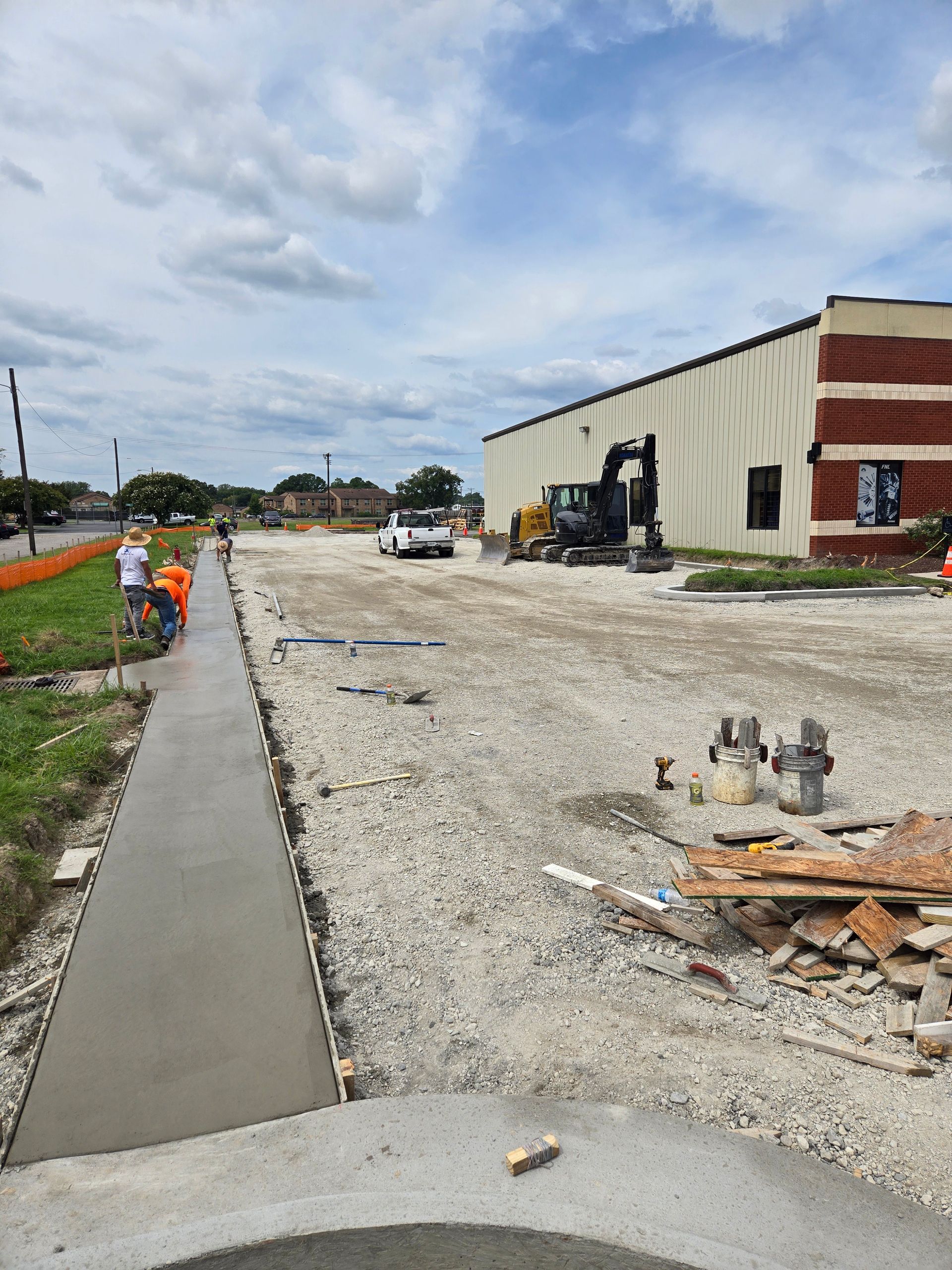 Construction workers install a new concrete sidewalk alongside a building under renovation with construction equipment.