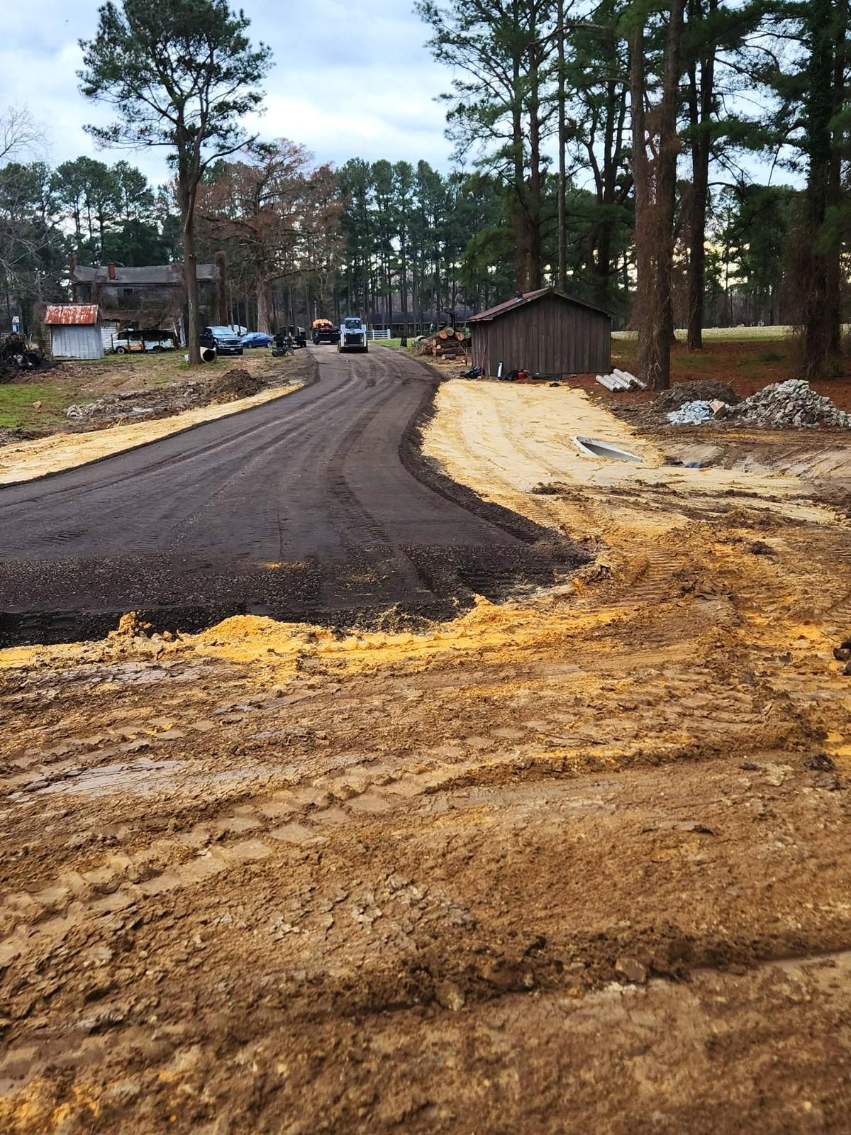 A newly paved dark asphalt road curves through a rural, wooded area with unfinished dirt shoulders and a small shed nearby.