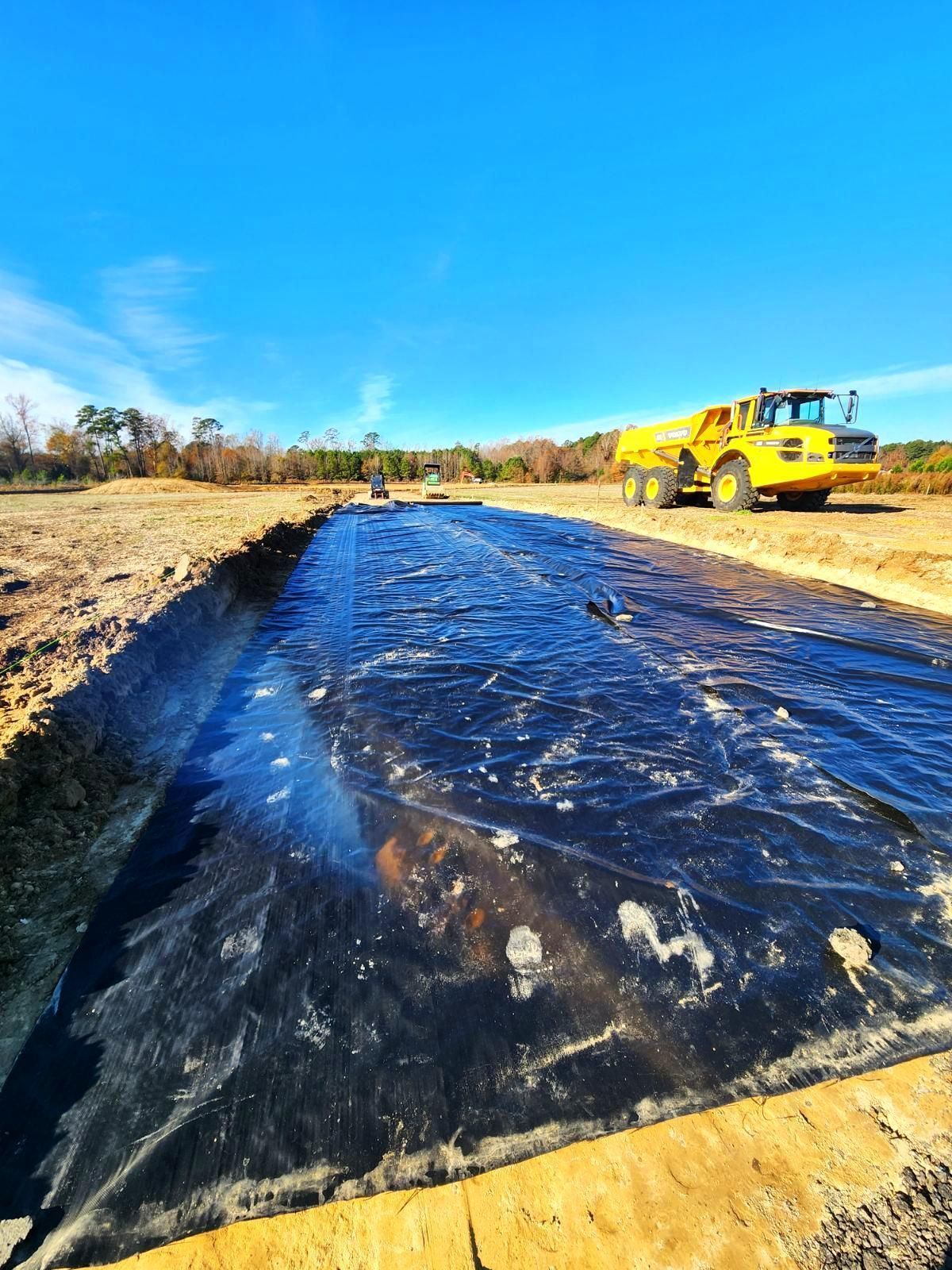 A yellow articulated dump truck sits beside a trench lined with a large, black plastic sheet under a clear blue sky.