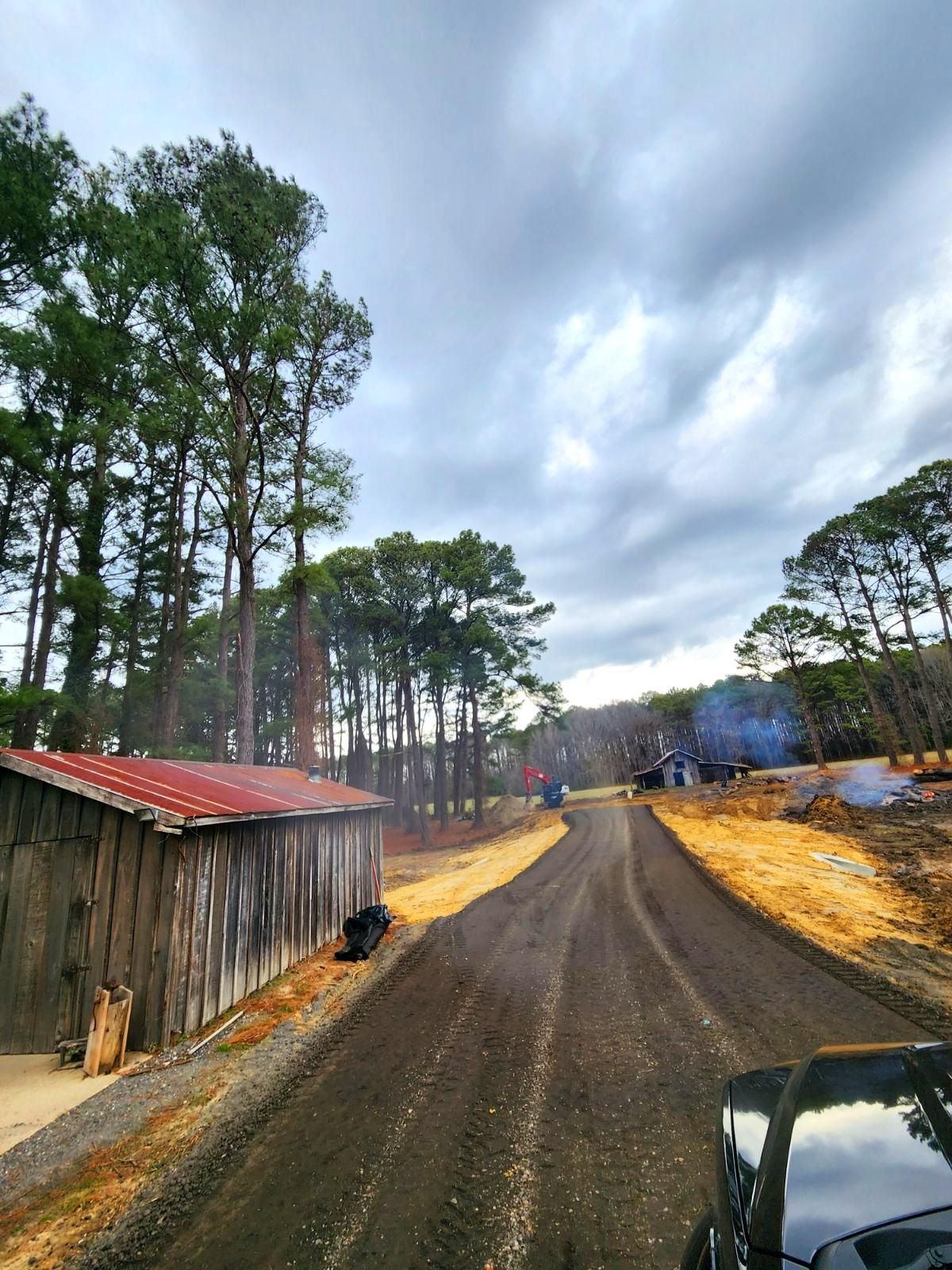 A gravel road leads past a rustic wooden shed toward a forest under a cloudy, overcast sky.