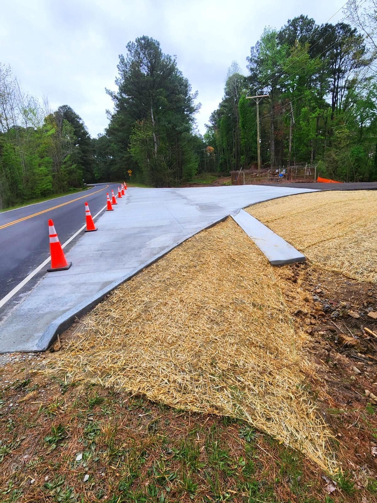 A newly paved concrete roadside apron and curb transition leading to a wooded road, lined with orange safety cones.