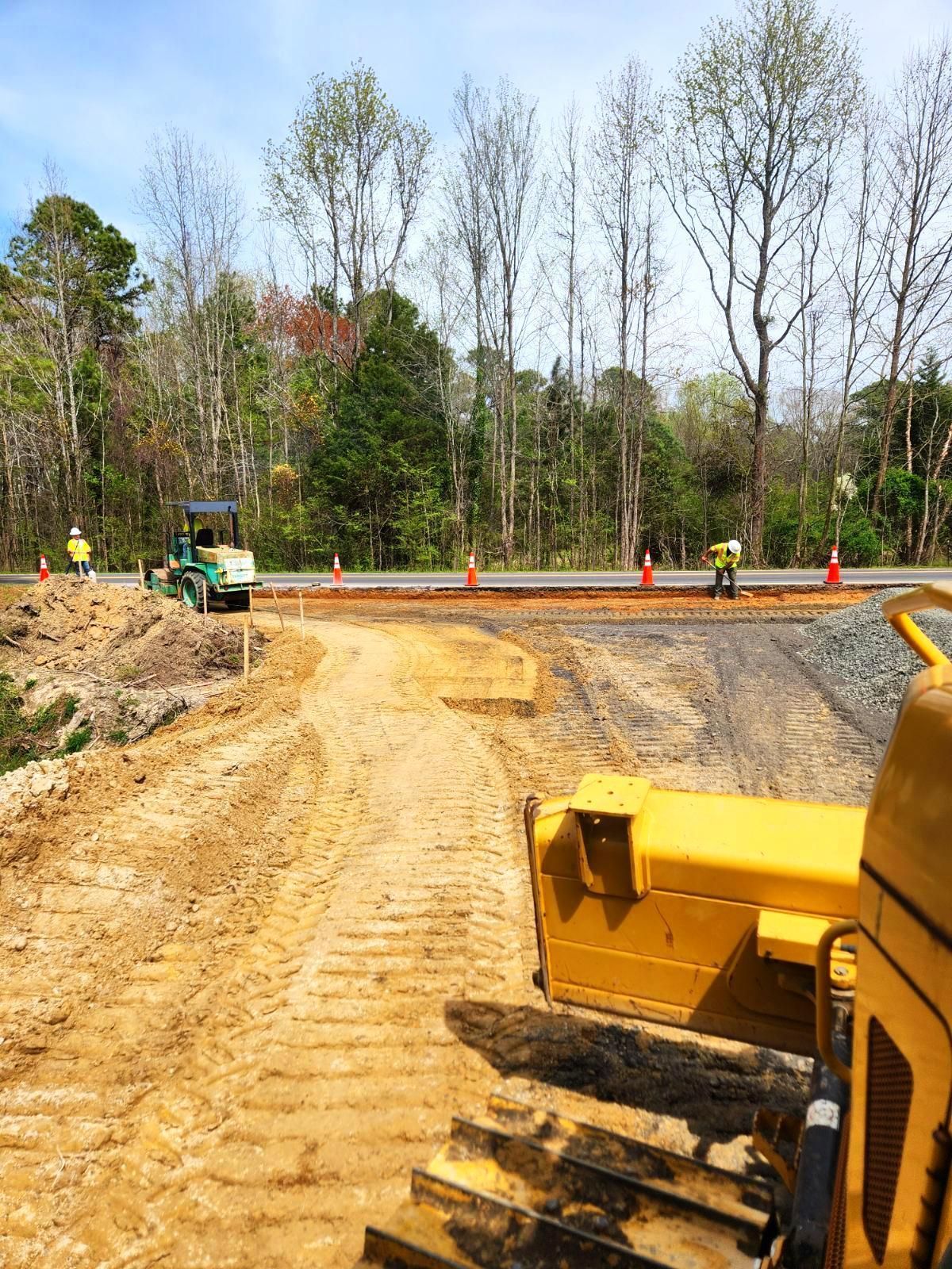 A yellow bulldozer in the foreground of a sunny construction site, with workers and orange cones near a paved road.