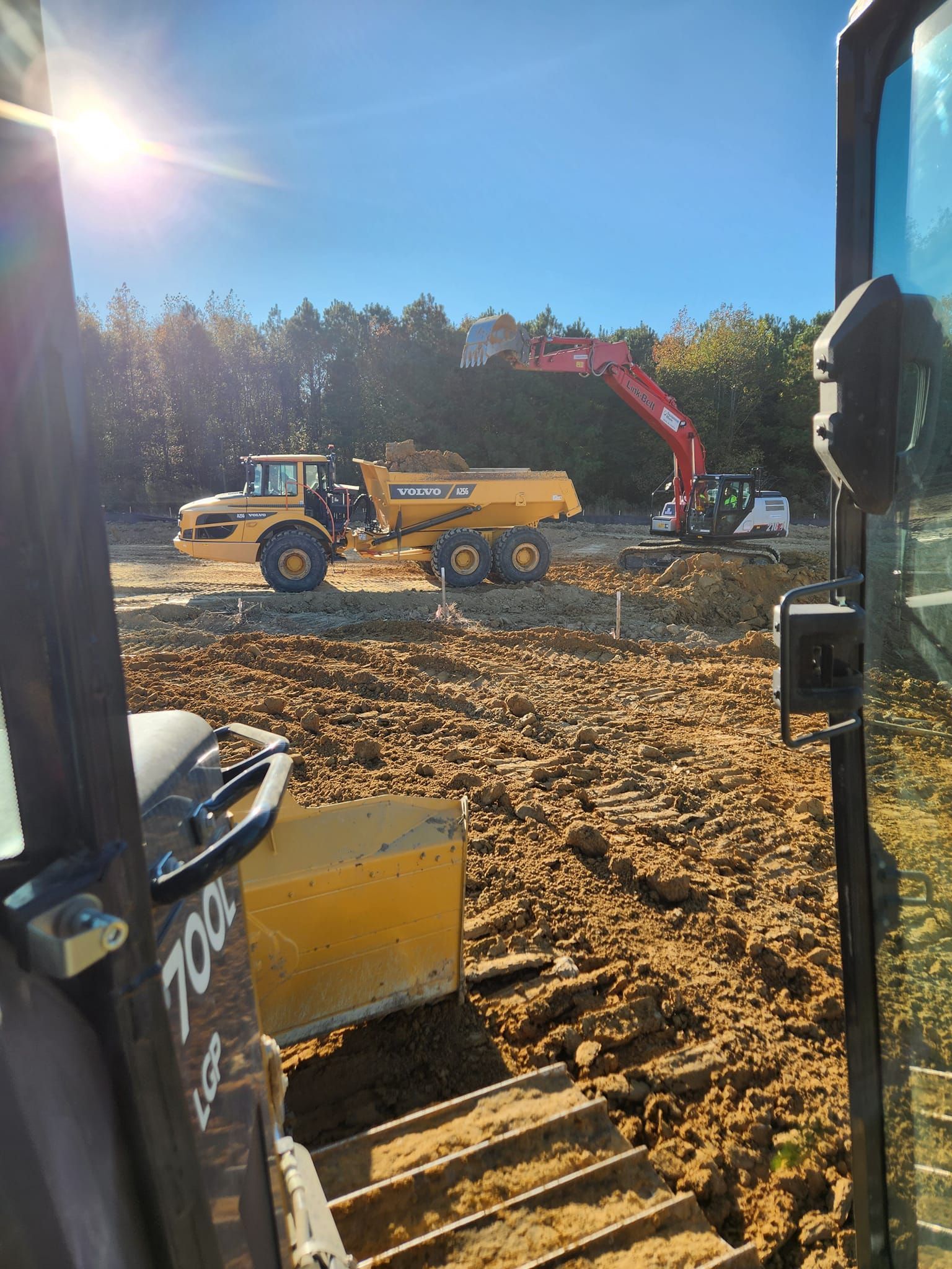 A view from a construction vehicle cab showing a yellow dump truck being loaded by an excavator on a dirt site.