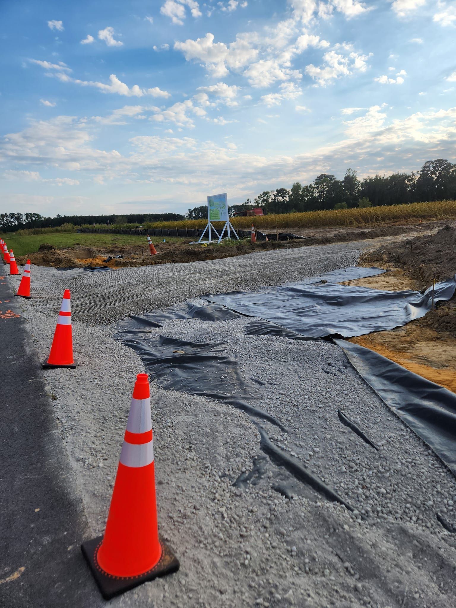Traffic cones line a gravel construction entrance with exposed black landscaping fabric on a sunny day.