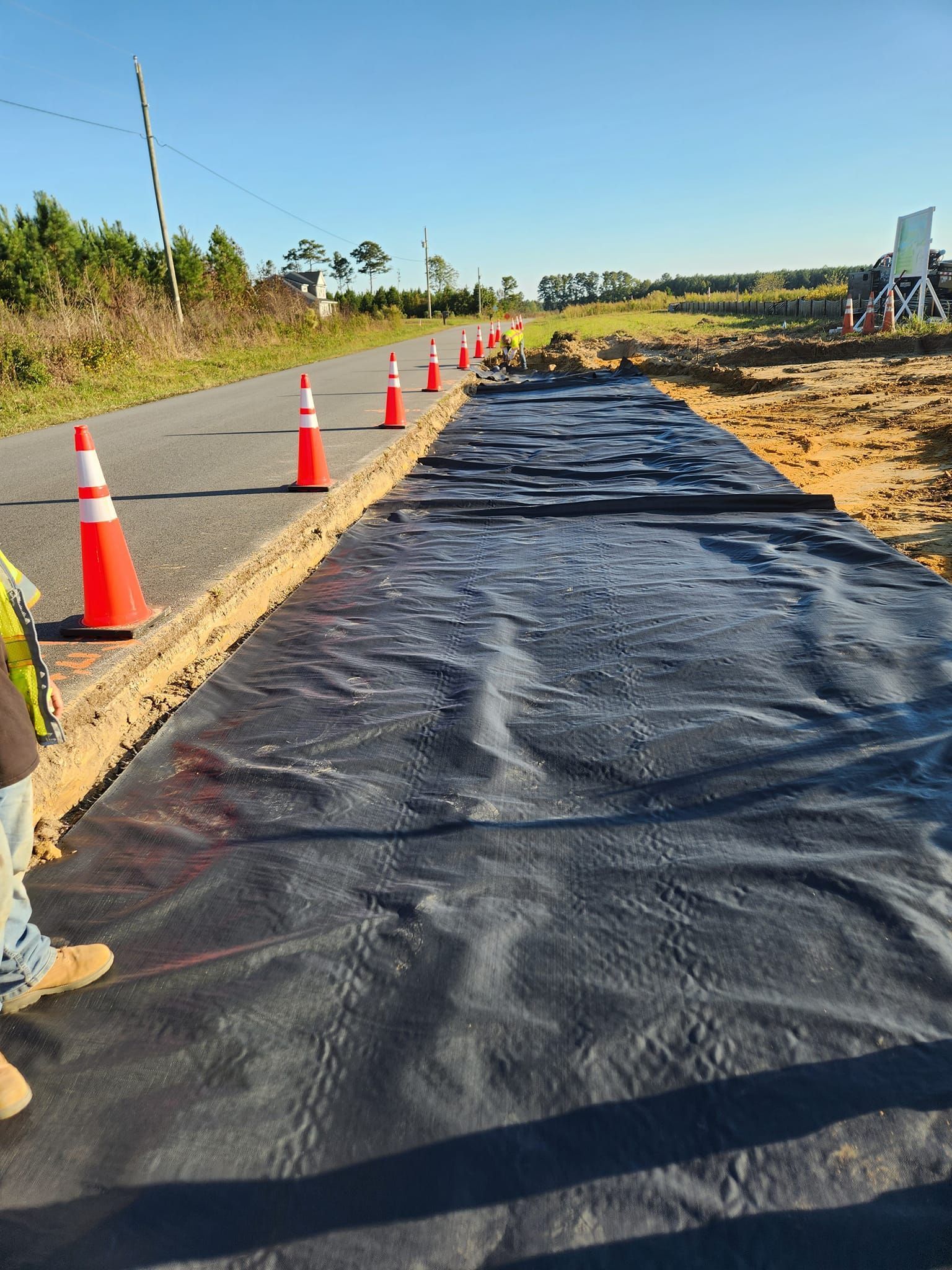 Black construction fabric laid out along a roadside, bordered by a line of orange traffic cones and a dirt excavation area.