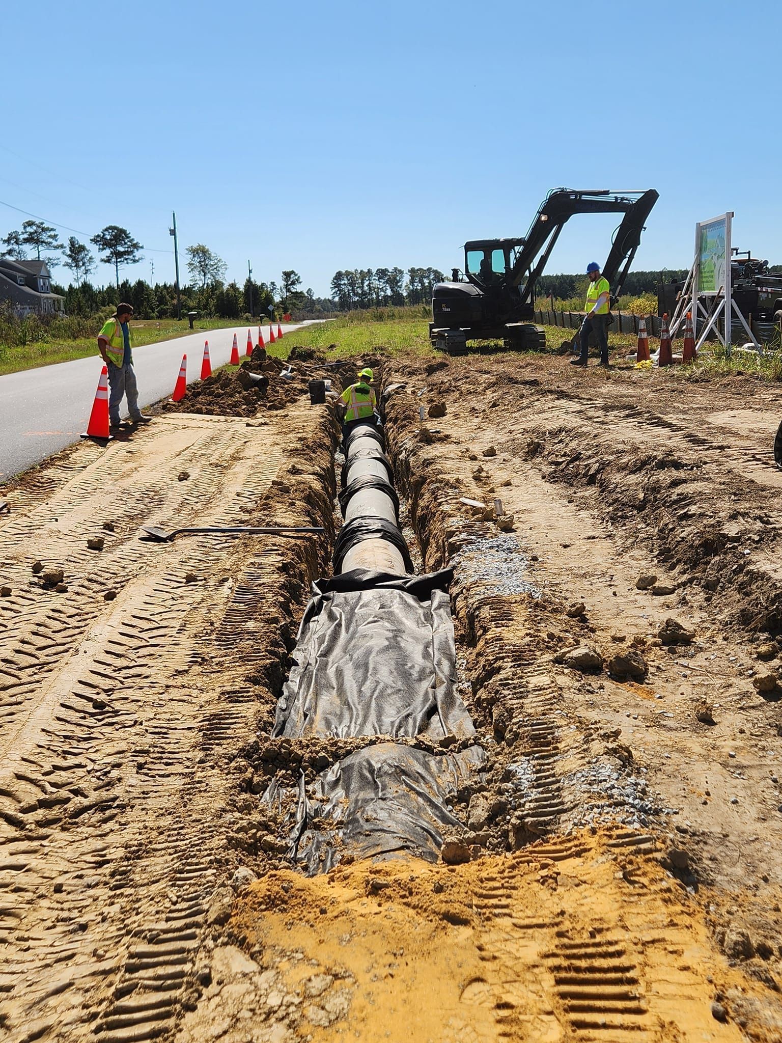 Construction workers install a pipe in a trench beside a road, with an excavator nearby on a sunny day.