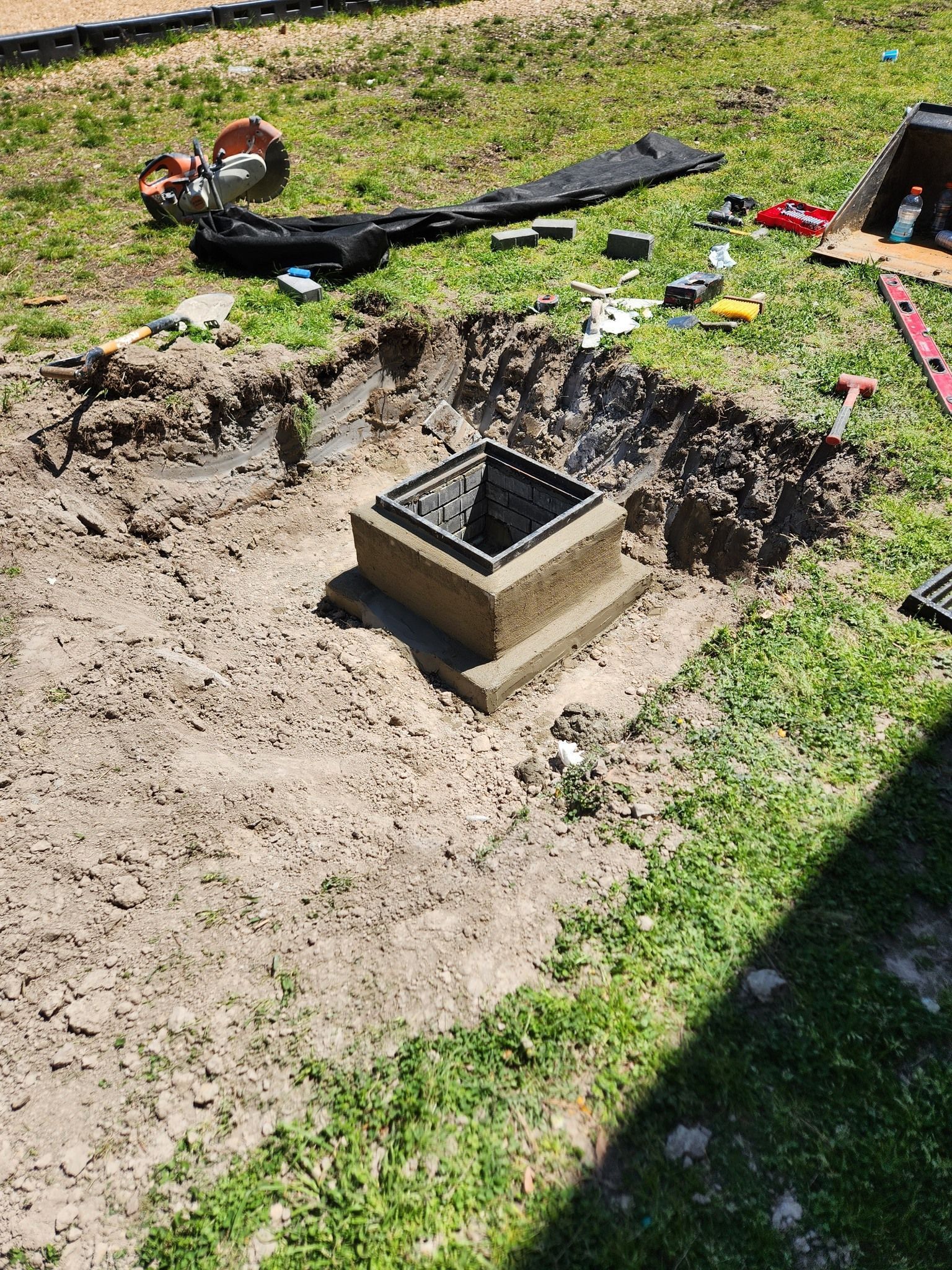 A square drainage basin set in a dug-out patch of grass and dirt with construction tools nearby on the lawn.
