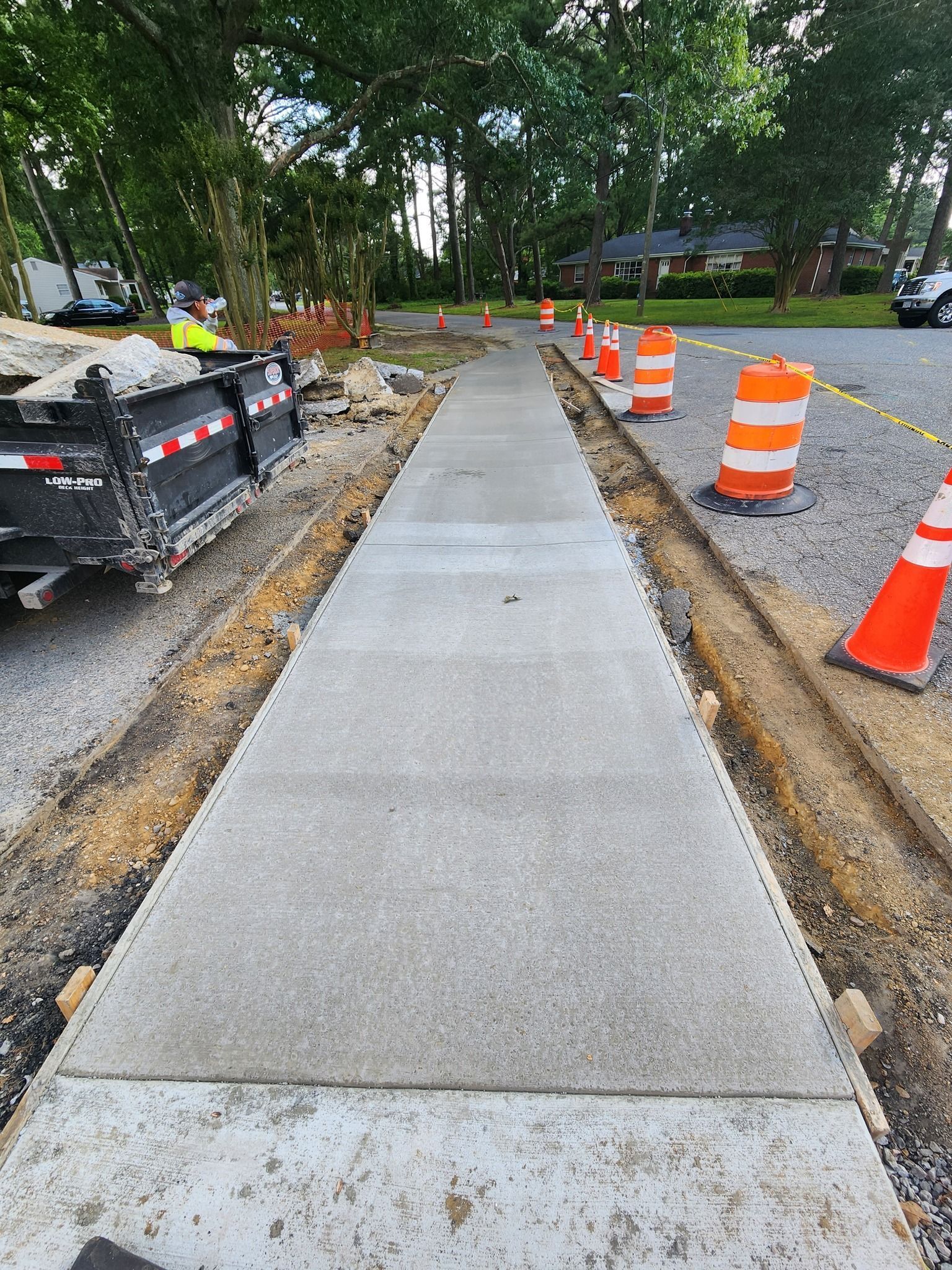 Freshly poured concrete sidewalk under construction, lined with orange safety barrels on a gravel shoulder.