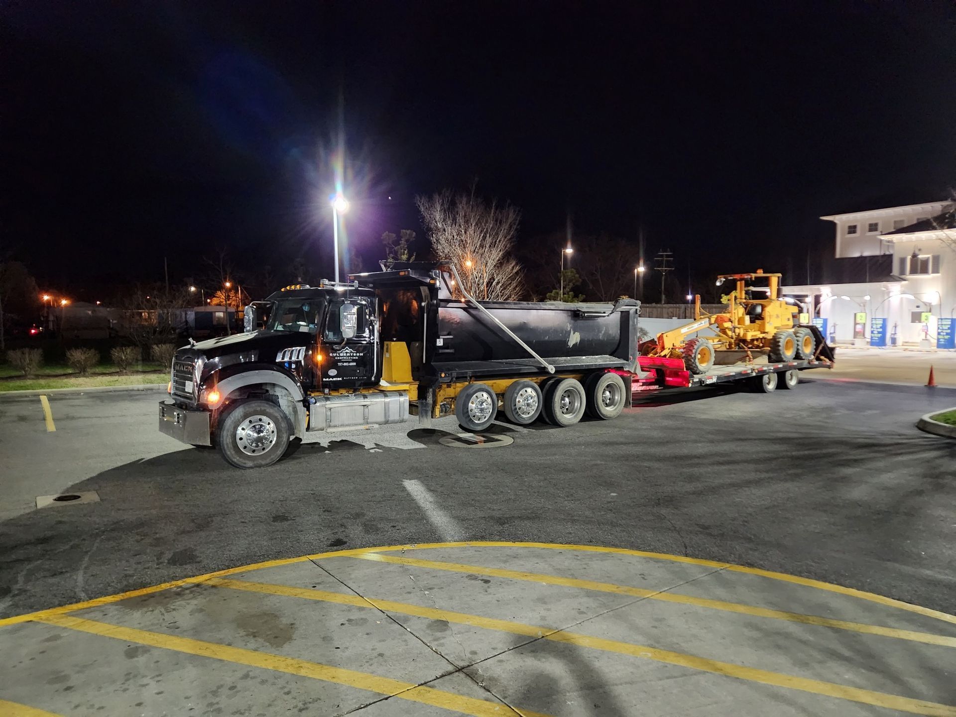 A black dump truck tows a flatbed trailer carrying a yellow construction grader in a parking lot at night.