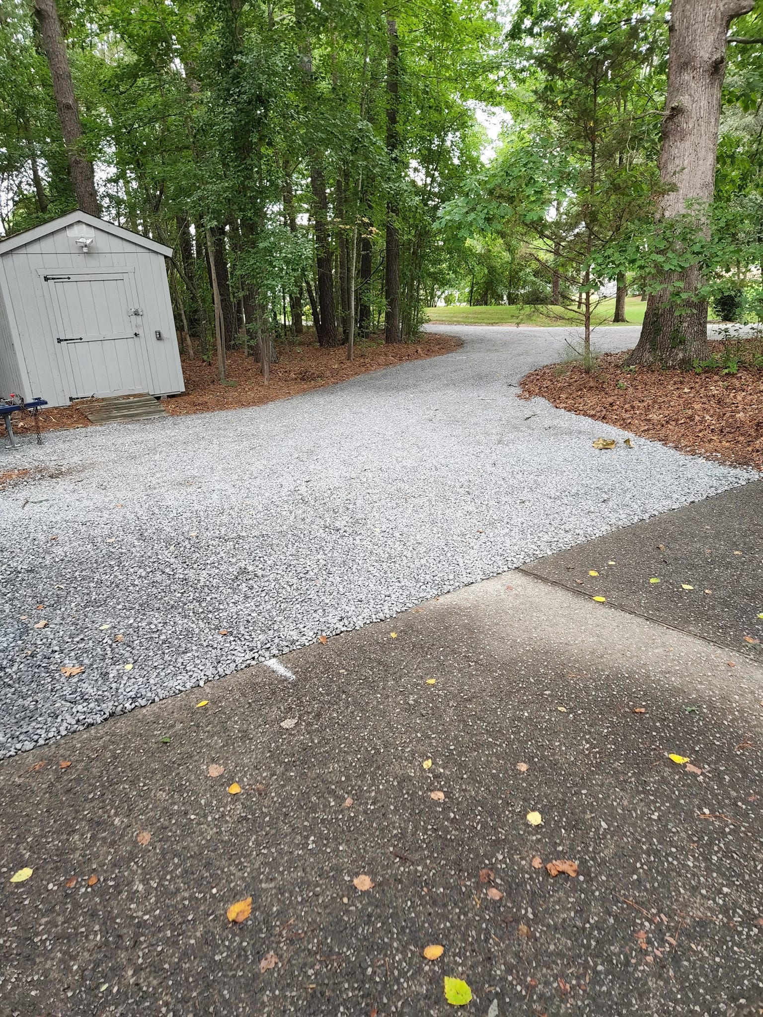 A gravel driveway transitions into a paved asphalt path next to a small storage shed, with trees in the background.