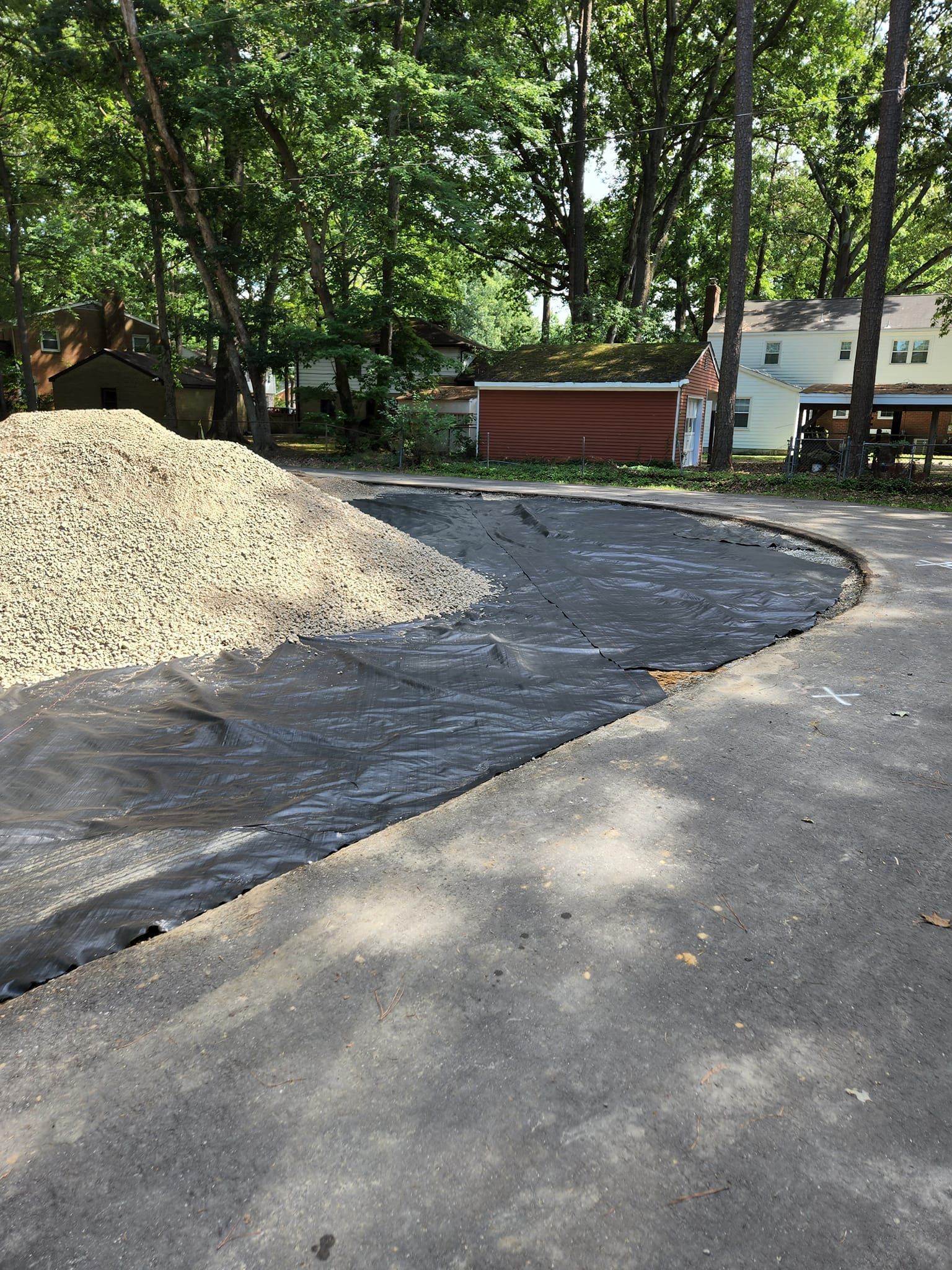 A large pile of gravel sits on black landscape fabric covering a portion of a driveway in a residential yard.