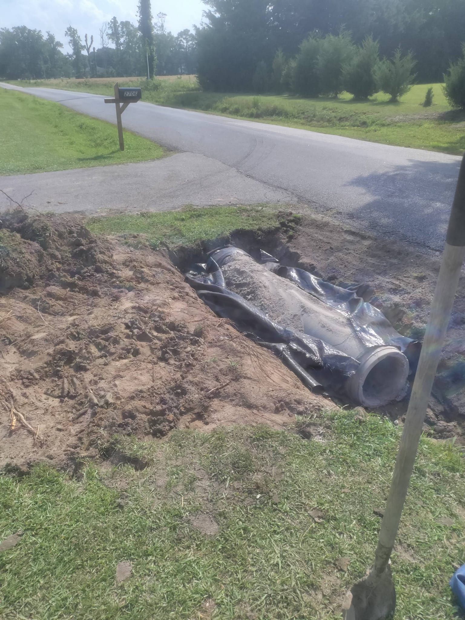 A drainage pipe sits in a trench beside a rural road, partially wrapped in black plastic, with a shovel in the foreground.