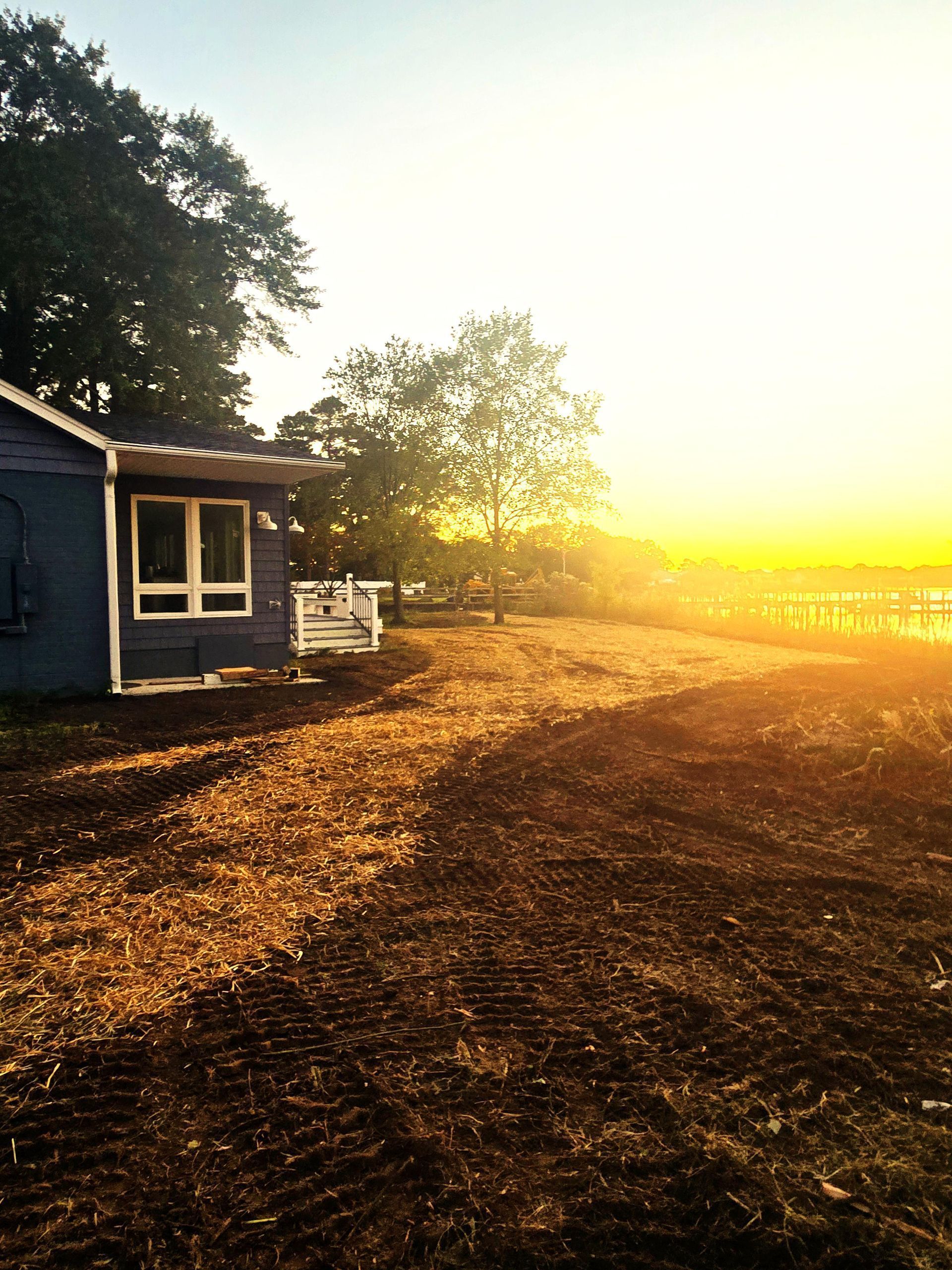 A blue cottage sits by a waterfront at sunset, casting a warm golden glow over a recently cleared, dirt-covered yard.