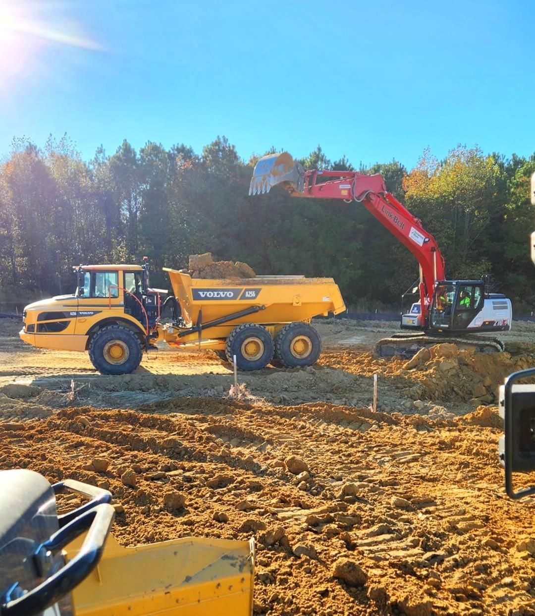 A red excavator loads dirt into a yellow Volvo articulated dump truck on a sunny, dirt-filled construction site.