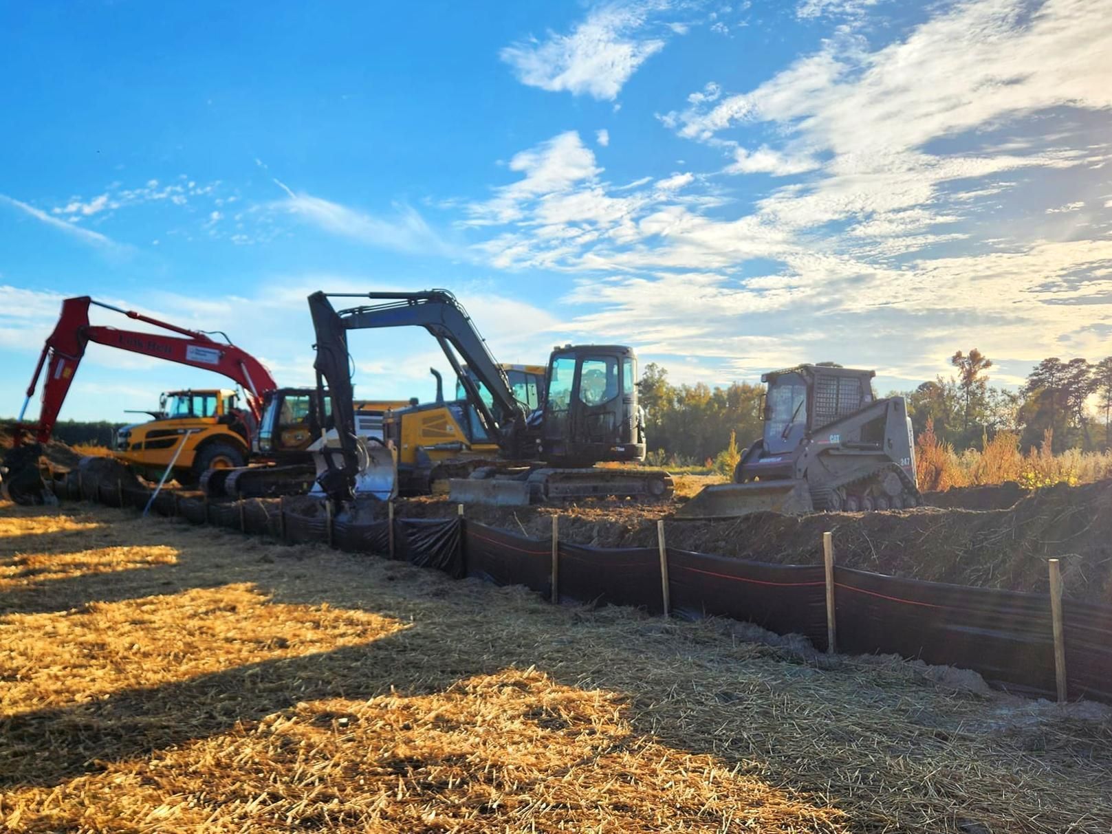 Three construction vehicles, including two excavators and a skid steer, parked at a job site with a black silt fence.