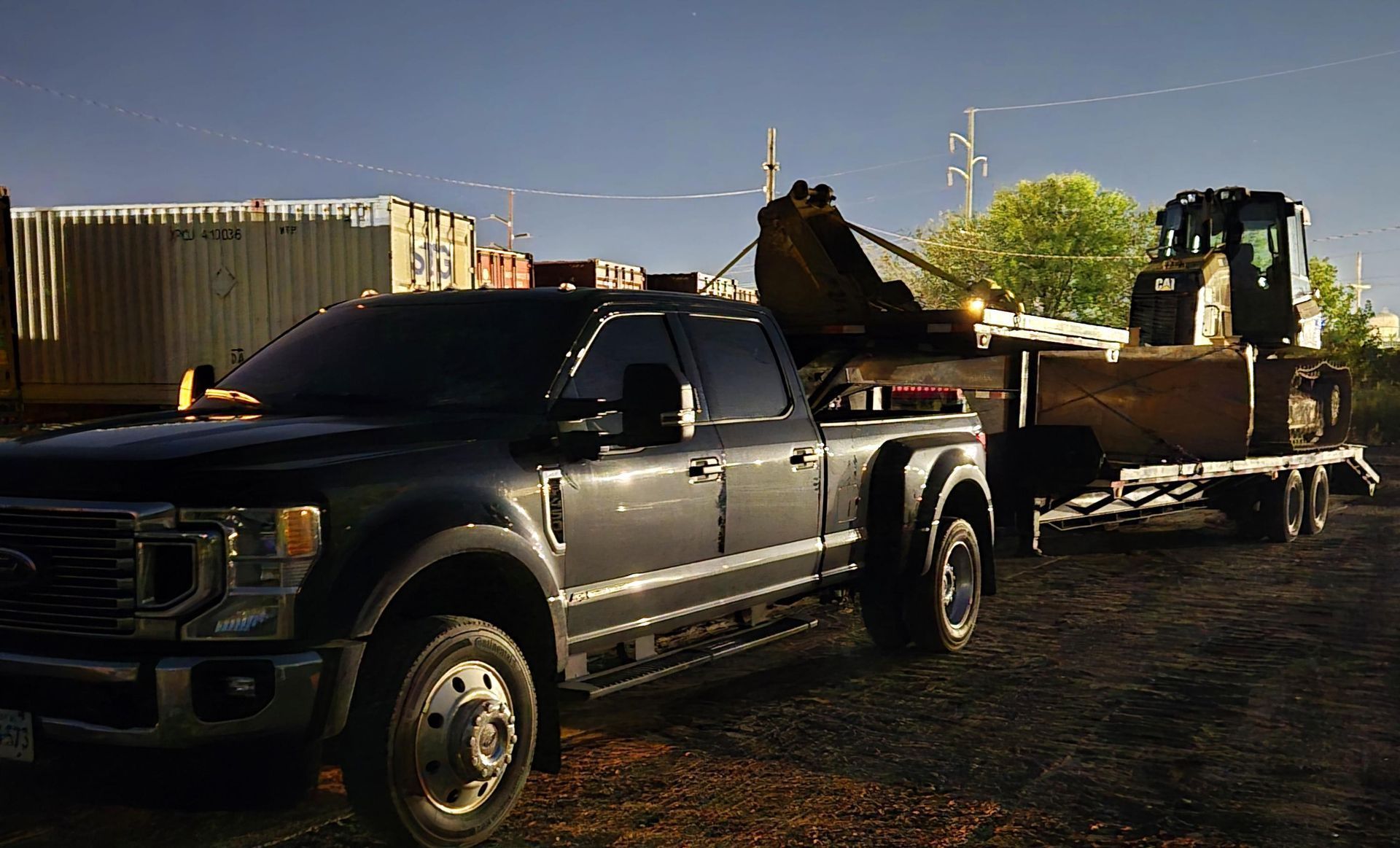 A dark-colored dually pickup truck towing a flatbed trailer with heavy construction machinery on a dirt lot.