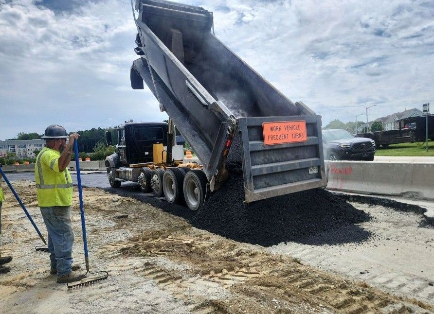 A construction worker in a high-visibility vest stands near a dump truck depositing asphalt on a road construction site.