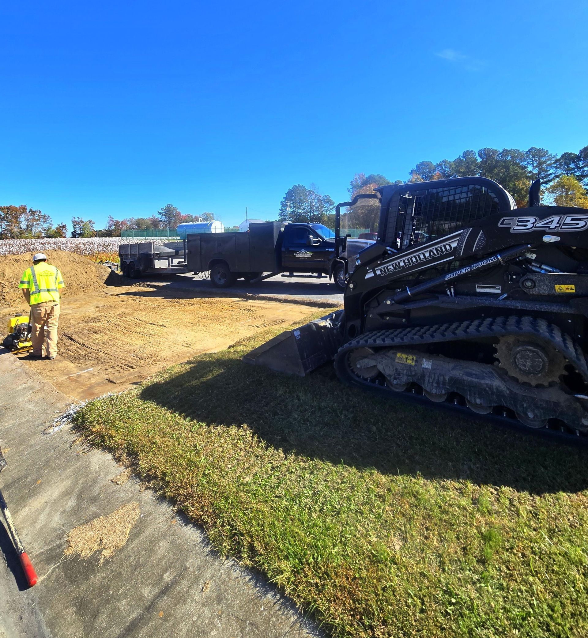 A worker in a high-visibility yellow vest stands near a tracked skid steer loader at a sunny construction site.
