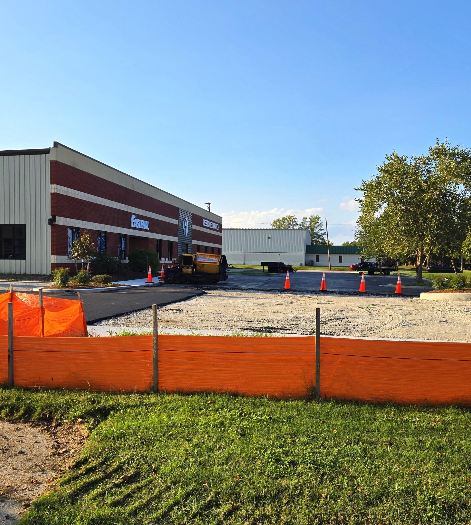 A red and white industrial building stands behind an orange safety fence and gravel construction site under a clear sky.