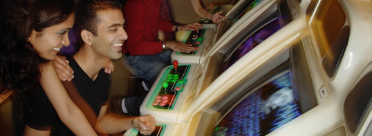People smile while playing side-by-side retro arcade games in a dimly lit setting.