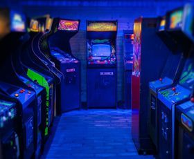 A dimly lit arcade room with rows of vintage video game cabinets under glowing blue light.