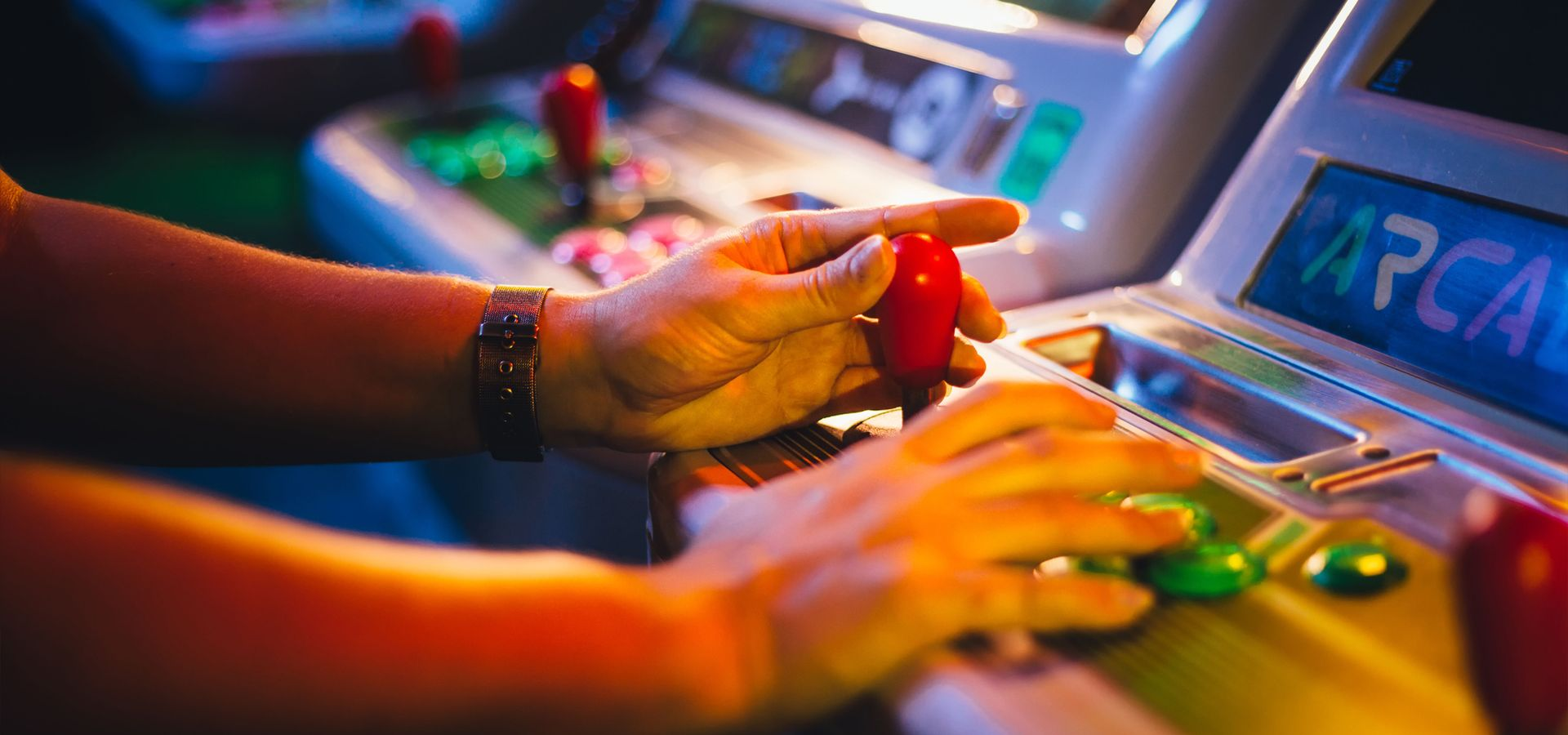 Close-up of hands playing a video game at an arcade machine, using a red joystick and a grid of colorful buttons.