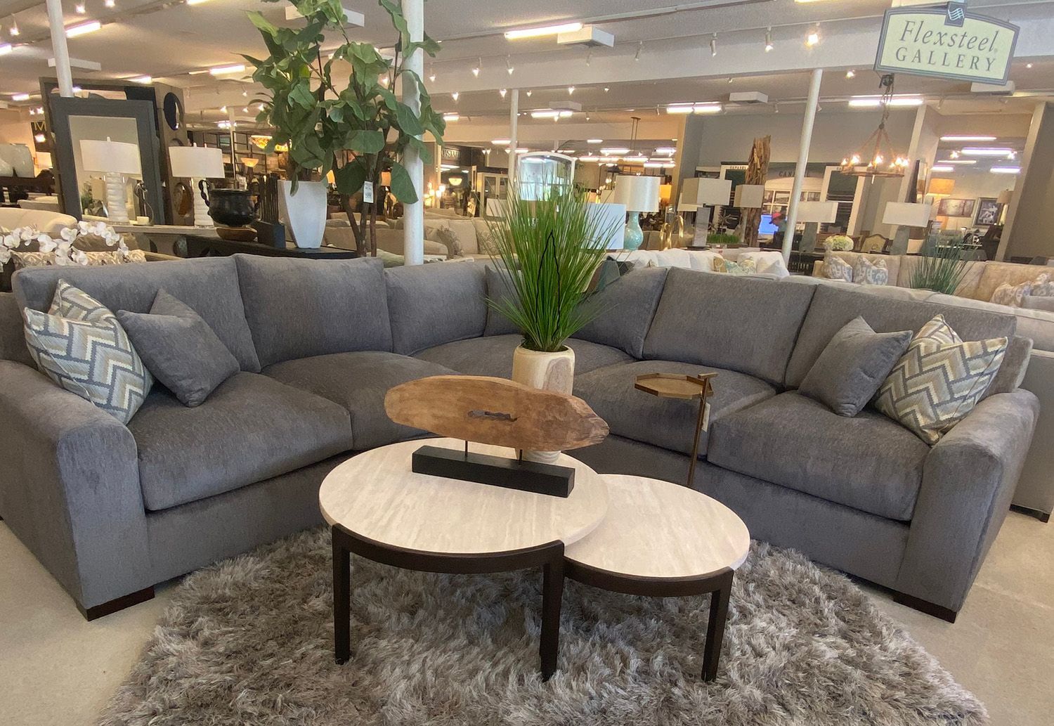 Gray sectional sofa and two-tone coffee tables on a fluffy rug in a furniture store.