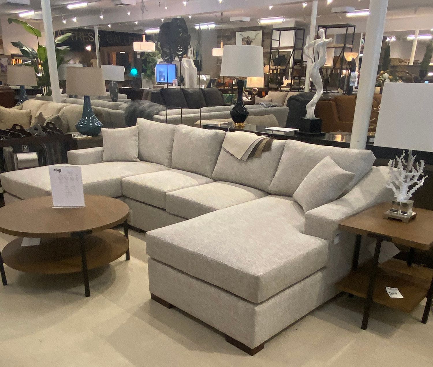 A spacious light-colored sectional sofa with a round coffee table and side table in a furniture store.
