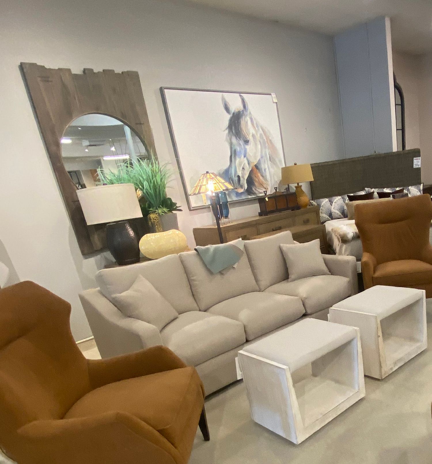 Living room display: beige sofa, two brown chairs, white ottomans, horse painting, mirror.
