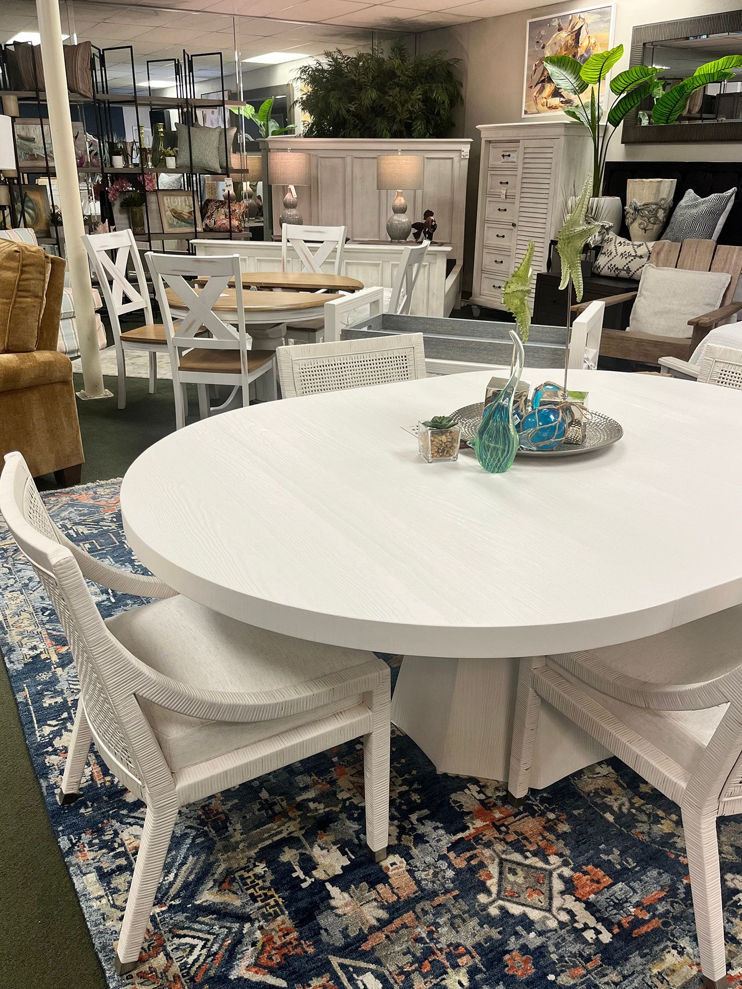 White oval dining table with woven chairs on blue patterned rug, furniture store.