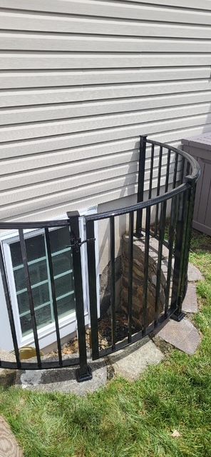Curved black metal window guard beside a basement window on beige siding, above grass and concrete blocks