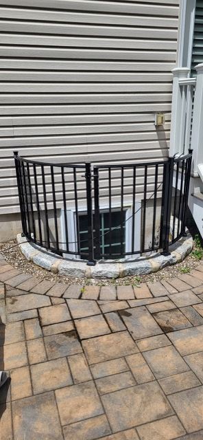 Curved black metal railings around a basement window well beside beige siding and stone pavers