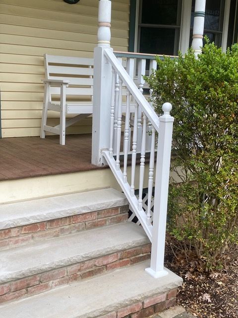 Front porch steps with a white railing, chair, and green bushes beside a house