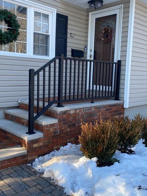 Front porch with black railing, brick steps, wreaths, and snow on the ground