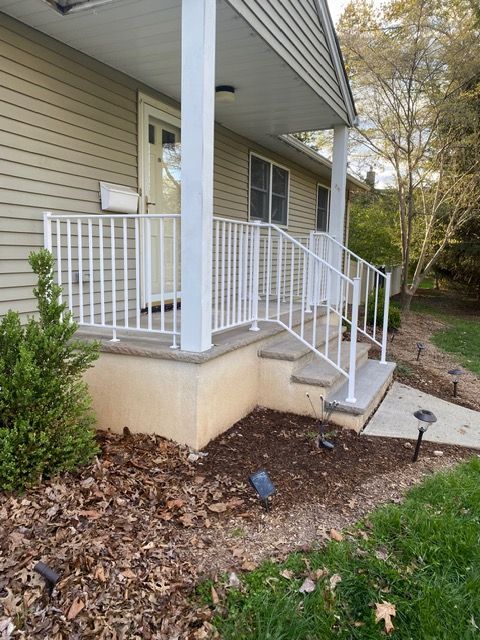 Front porch with white railings and steps leading to a beige house, bordered by mulch and shrubs.