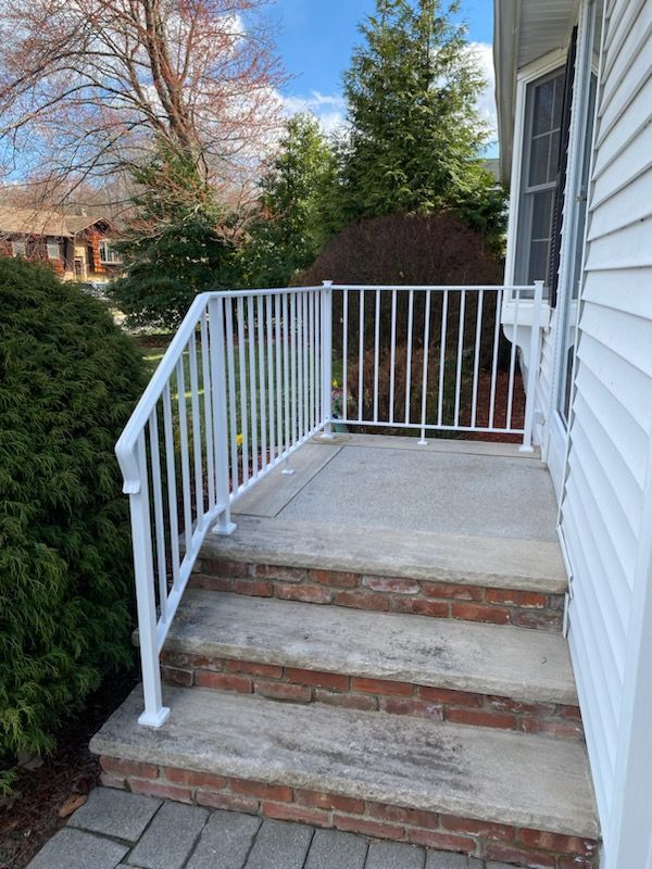 Front steps to a house with brick risers and white railings beside a driveway and shrubs