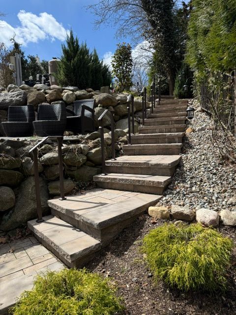 Stone steps in a landscaped garden with rock walls, potted plants, and trees under a blue sky